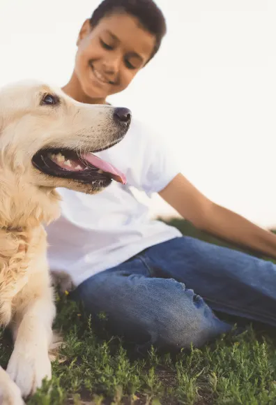 Little boy sitting on grass with his dog laying down next to him as he pets the top of his dogs head. 